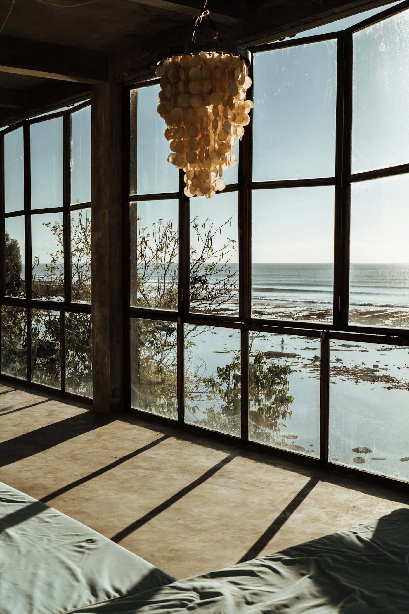Sala de casa de praia com janelões de vidro de frente para o mar e luminária de conchas