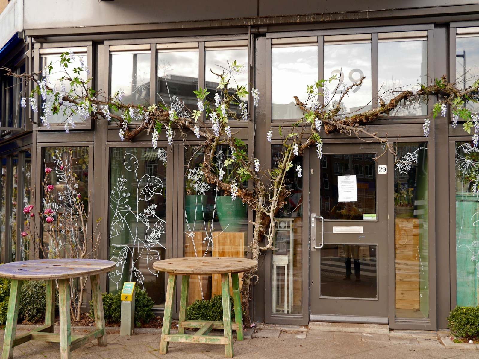 Fachada de comércio com portas e vitrines de vidro, mesas na calçada e plantas