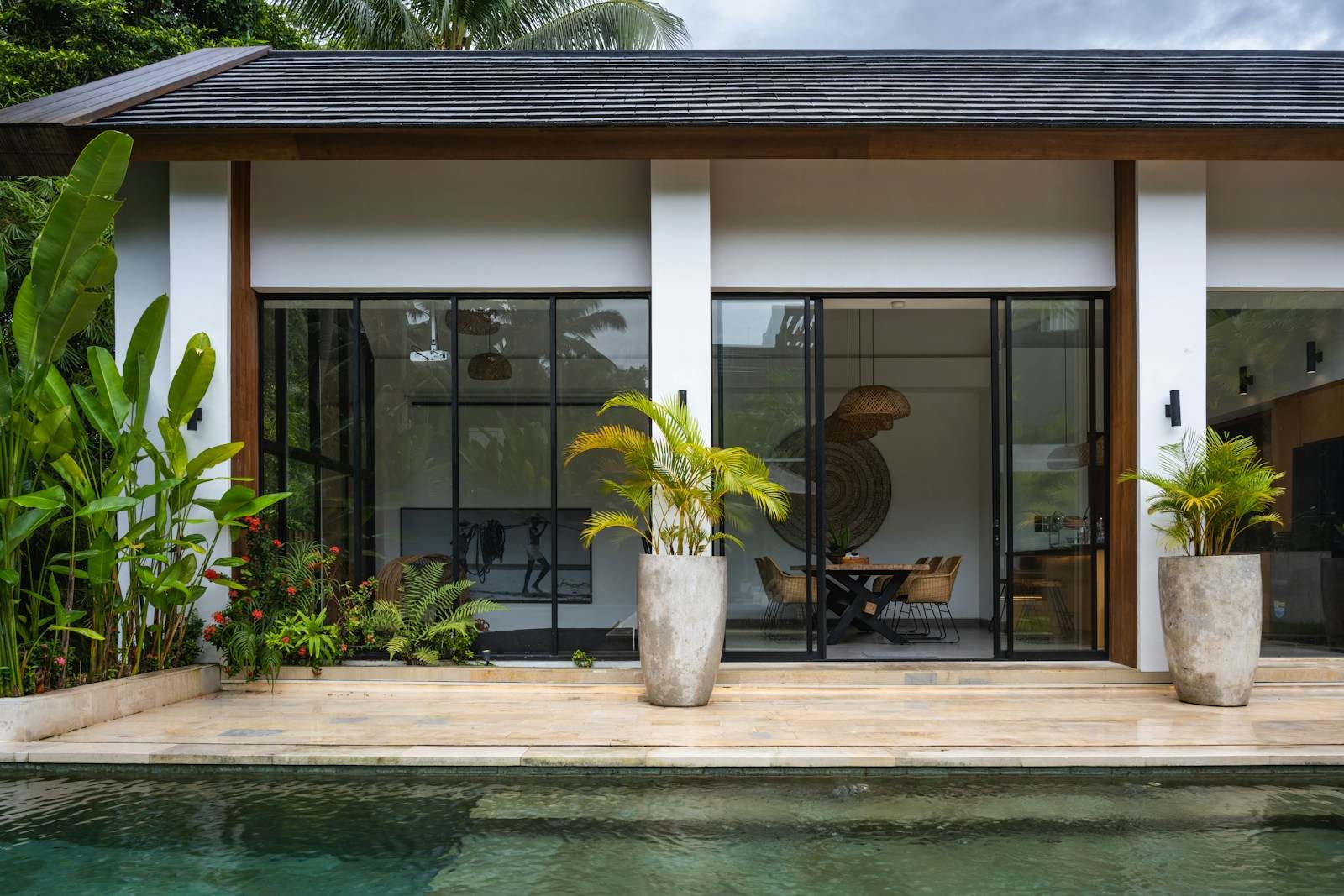 Casa térrea de praia com portas de vidro abertas para a piscina, entre plantas tropicais